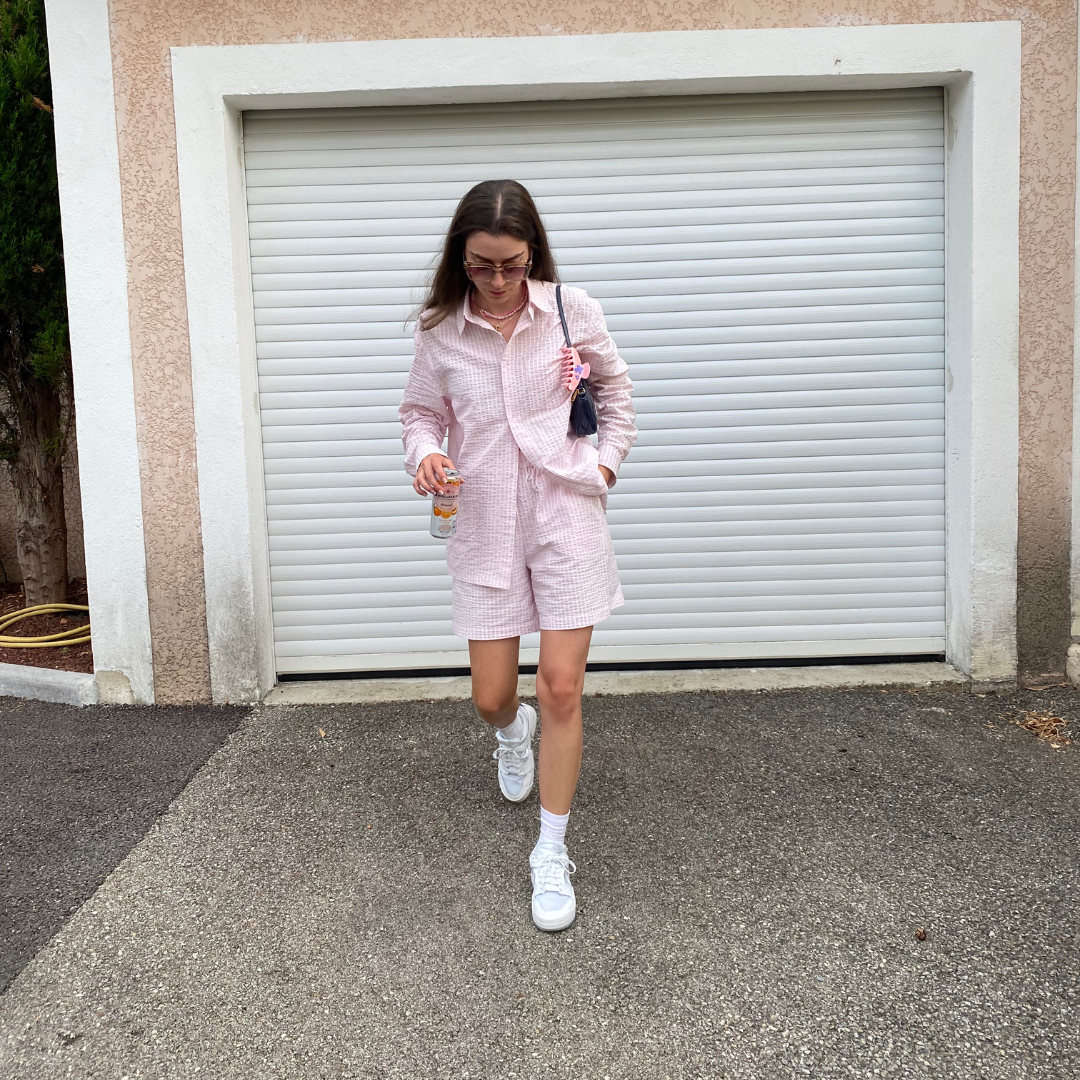 Eine Frau in einem rosa-weiß karierten Outfit geht auf einer gepflasterten Einfahrt vor einem geschlossenen weißen Garagentor, mit einem Getränk und einer kleinen Handtasche in der Hand, mit offenem Haar, Brille und weißen Turnschuhen.