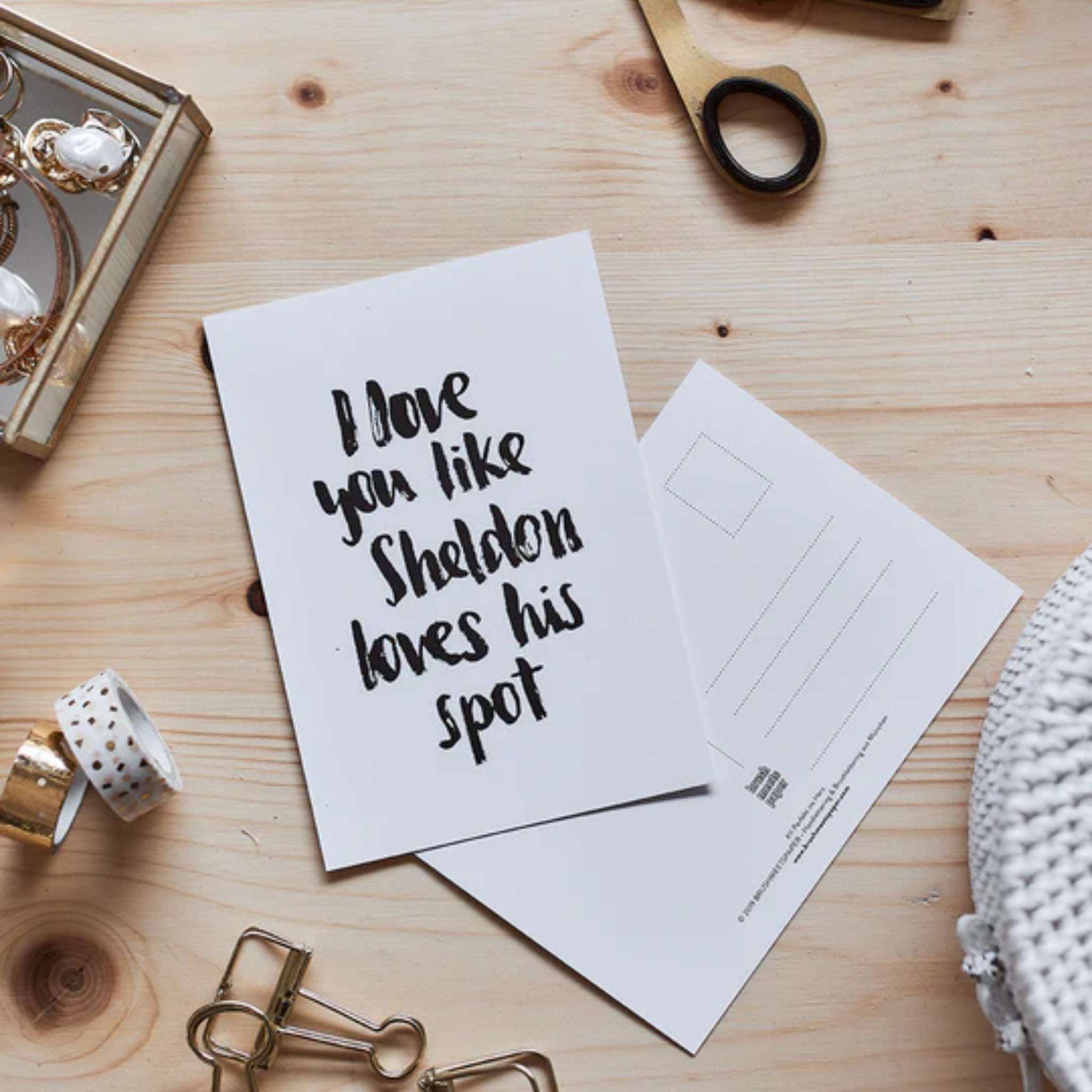 Eine Postkarte mit dem Text "I love you like Sheldon loves his spot" auf einem Holztisch, umgeben von Scheren und Band.