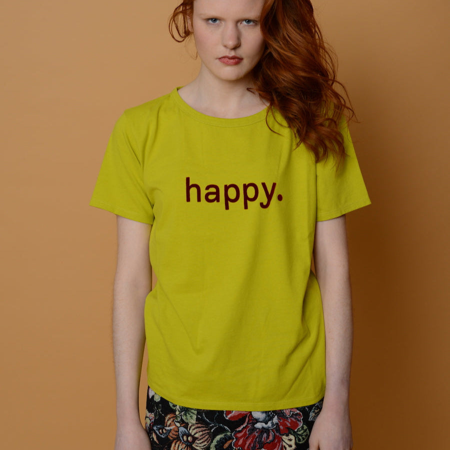 Frau mit rotem Haar trägt ein gelbes T-Shirt mit der Aufschrift "happy" und steht vor einem karamellfarbenen Hintergrund.