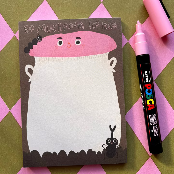 Notizblock in Pilzform mit der Aufschrift "So Mushroom for Ideas", daneben ein rosa Marker auf kariertem Untergrund.