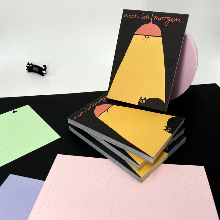 Notizblöcke mit der Aufschrift "mach ich morgen" und einem Katzenmotiv, umgeben von bunten Papieren.