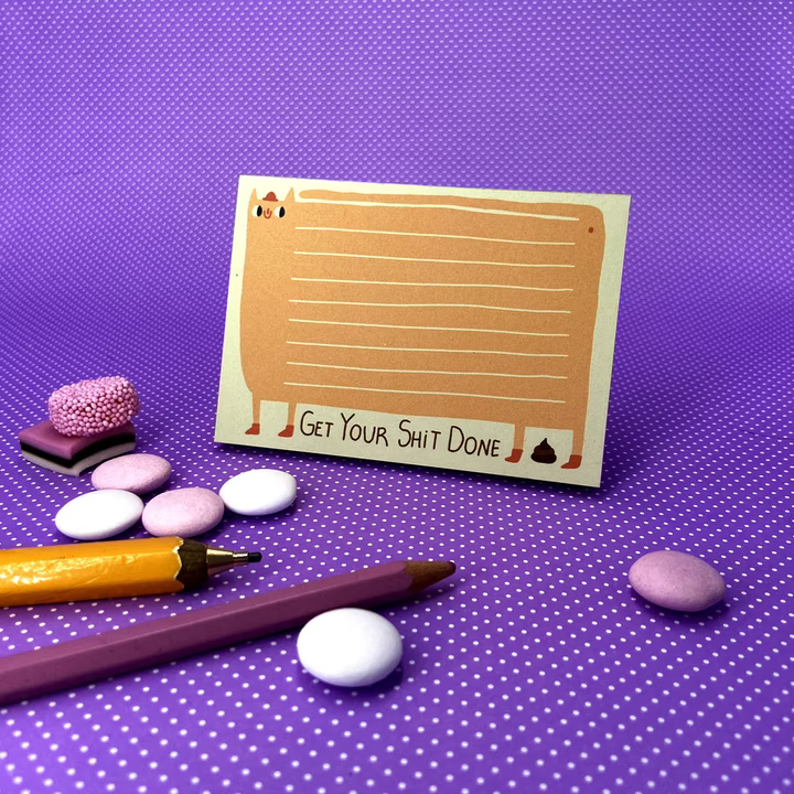 Notizkarte mit der Aufschrift „Get Your Shit Done“ liegt auf lila Punktmuster, umgeben von Stiften und Süßigkeiten.