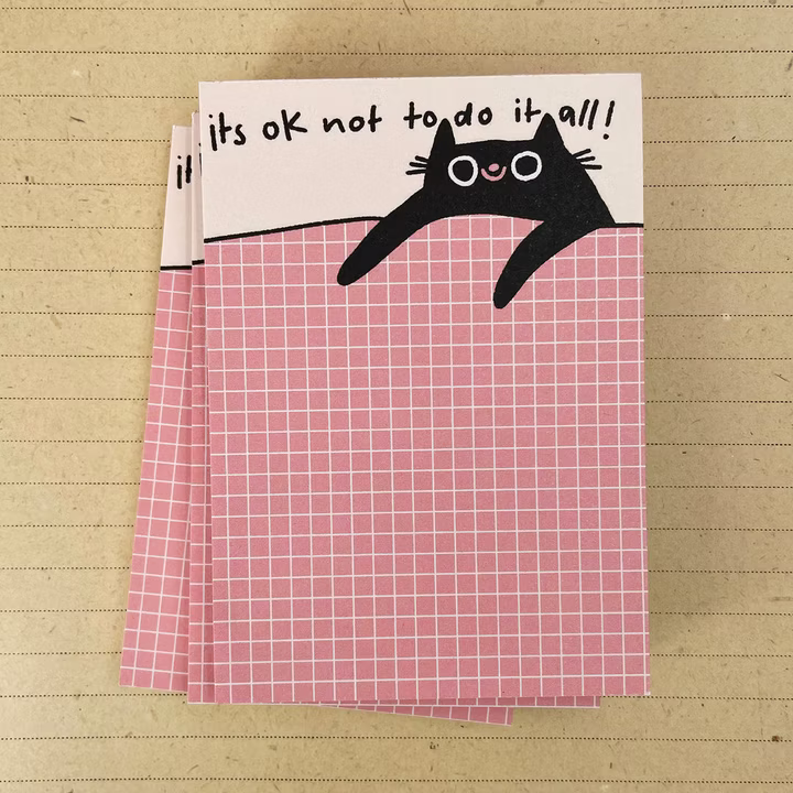 Notizblöcke mit einer gezeichneten Katze auf rosa Karos und dem Text "It's ok not to do it all!" oben.