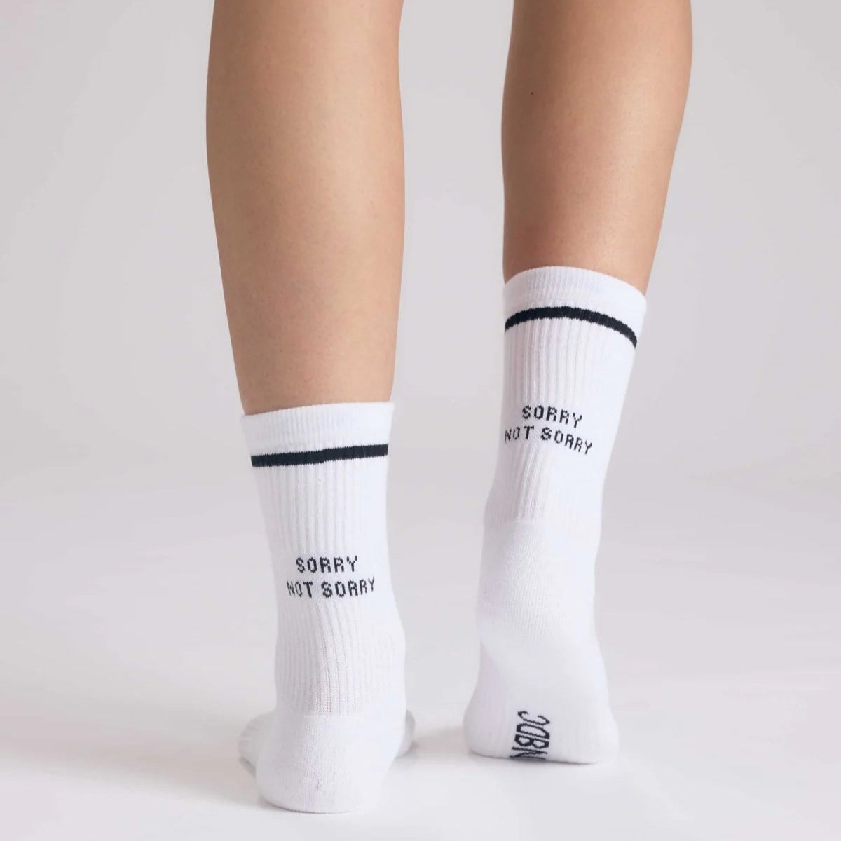 Eine Person steht mit gespreizten Füßen und trägt weiße Socken mit einem schwarzen Streifen und der Aufschrift SORRY NOT SORRY auf der Rückseite über den Fersen. Der Hintergrund ist einfarbig und hell gefärbt.