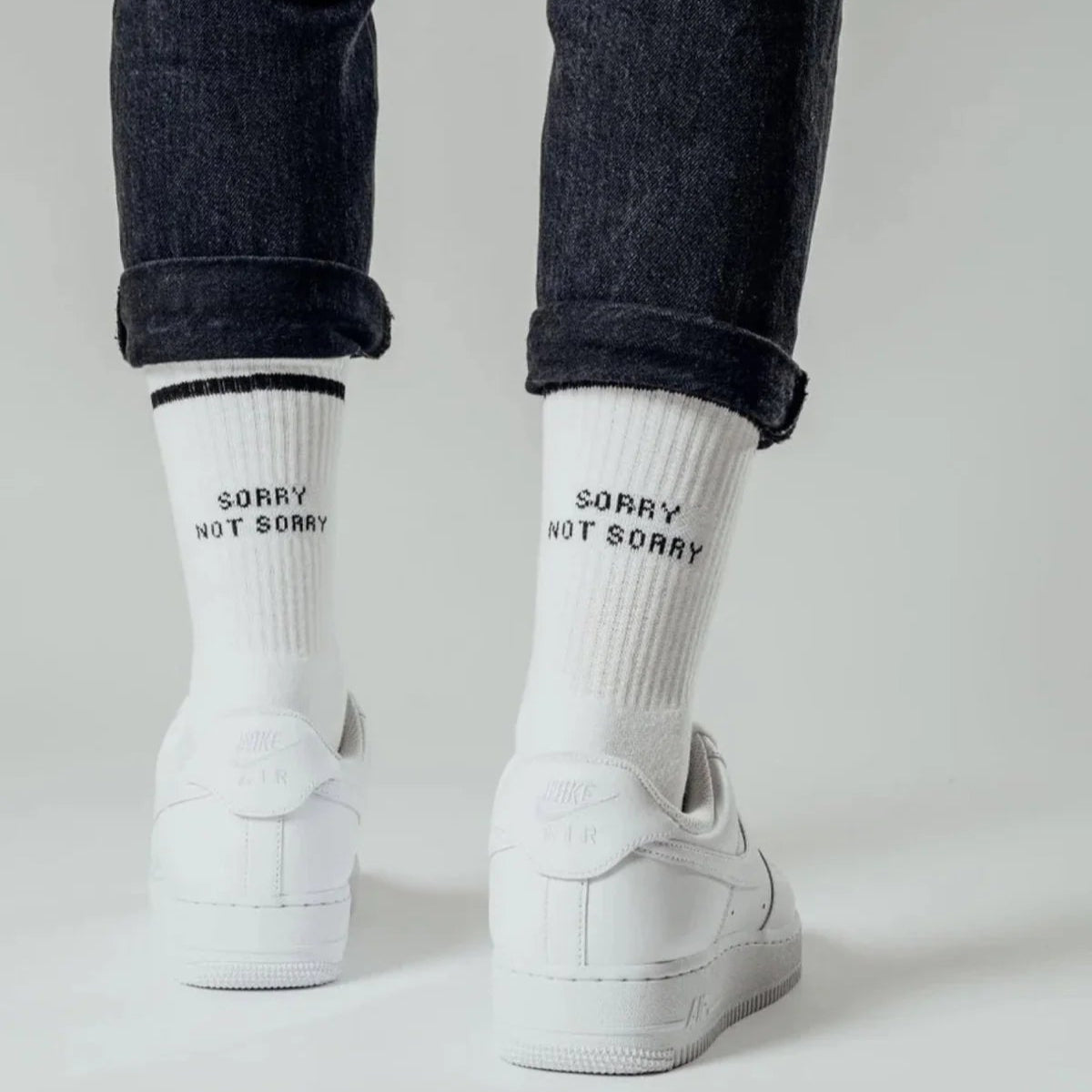 Eine Person in hochgekrempelten dunklen Jeans, weißen Turnschuhen und weißen Socken mit der Aufschrift SORRY NOT SORRY in schwarzer Schrift auf dem Rücken steht vor einem schlichten hellen Hintergrund.