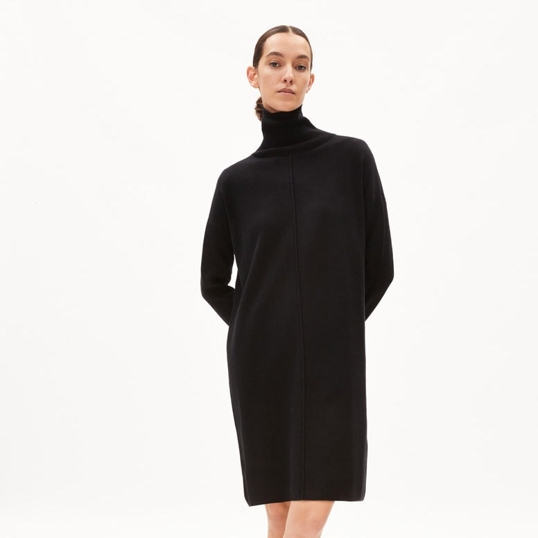 Eine Frau steht vor einem weißen Hintergrund und trägt ein schwarzes langärmeliges Kleid mit Rollkragen und Schuhe mit Leopardenmuster. Ihre Hände sind hinter dem Rücken verschränkt, und ihr Haar ist zurückgebunden.