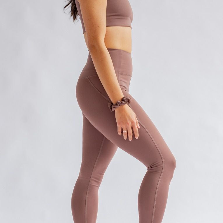 Eine Person in lilafarbenen Sportleggings und einem dazu passenden Sport-BH steht barfuß auf einem hellen Hintergrund. Sie wird von der Seite gezeigt, wobei ein Arm entspannt ist und das Gesicht aus dem Rahmen fällt.