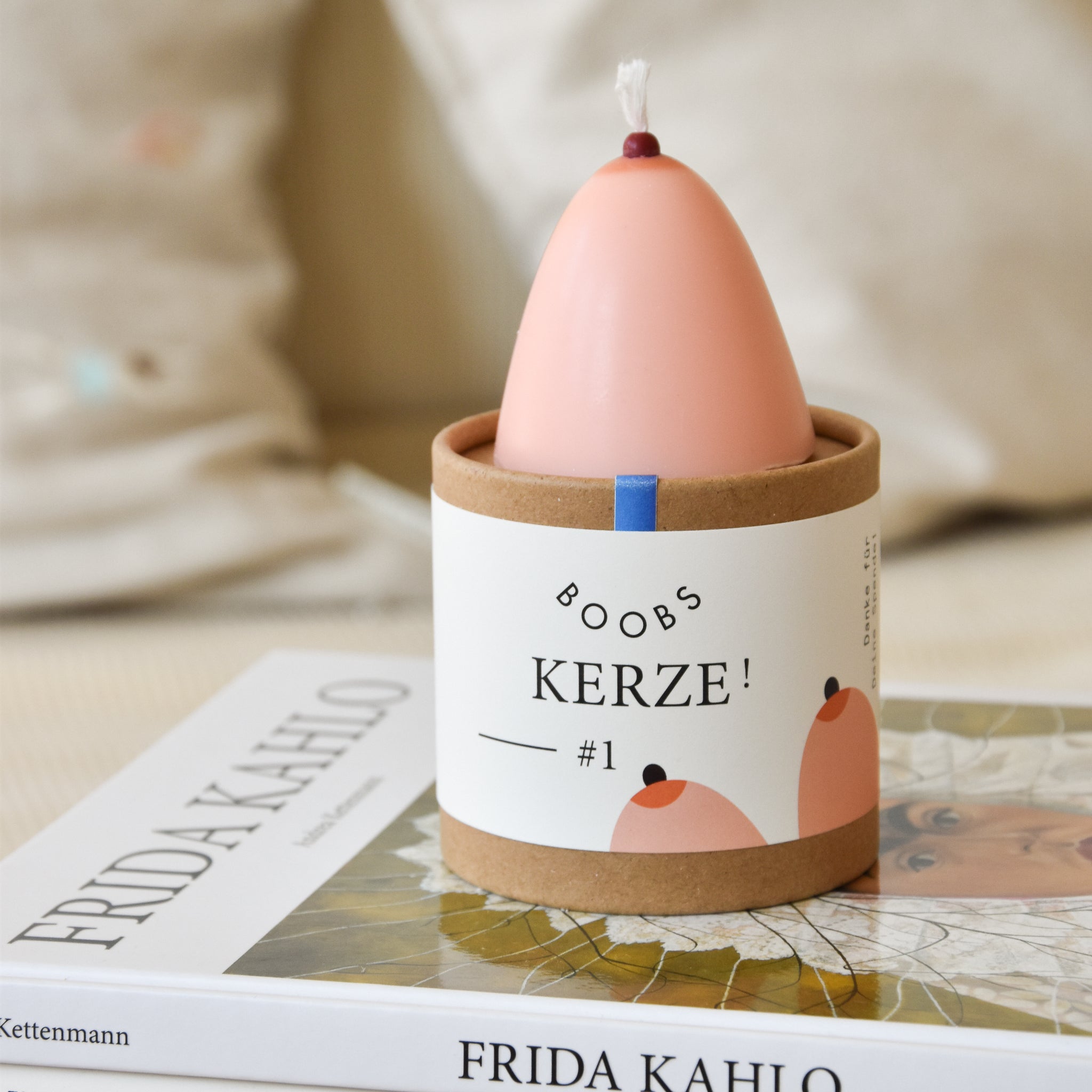 Eine Kerze in einer Verpackung beschriftet mit "Boobs Kerze #1", steht auf einem Frida Kahlo Buch.