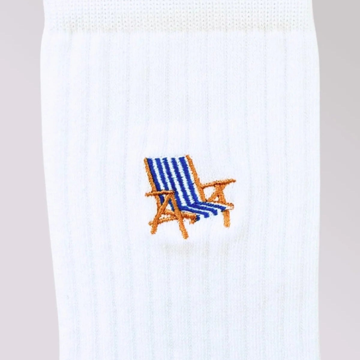 Eine Nahaufnahme einer weißen gerippten Socke mit einem kleinen gestickten blau-weiß gestreiften Strandkorb mit Holzarmlehnen.