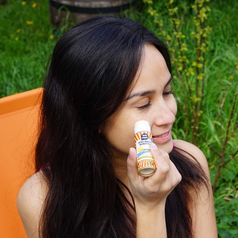 Eine Frau mit langen dunklen Haaren lächelt, während sie im Freien eine kleine Flasche Sonnencreme auf ihre Wange aufträgt, mit grünem Gras und Pflanzen im Hintergrund.