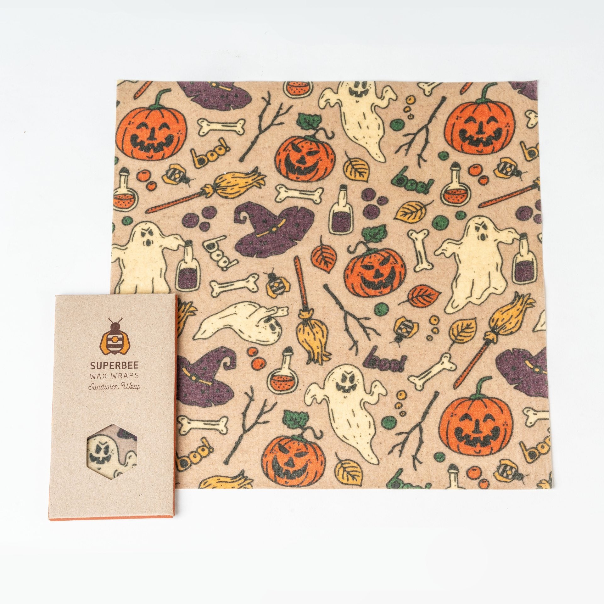 Eine quadratische Bienenwachsfolie mit einem Halloween-Muster, das Kürbisse, Gespenster, Knochen, Zaubertränke und Hexenhüte zeigt. Daneben befindet sich eine kleine Pappverpackung mit der Aufschrift SuperBee Wax Wraps auf weißem Hintergrund.