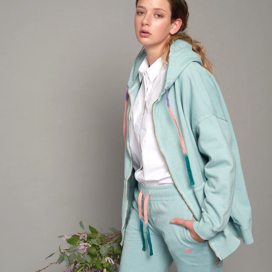 Frau in hellblauem Trainingsanzug und weißem Hemd steht vor grauem Hintergrund mit Blumenstrauß im Hintergrund.