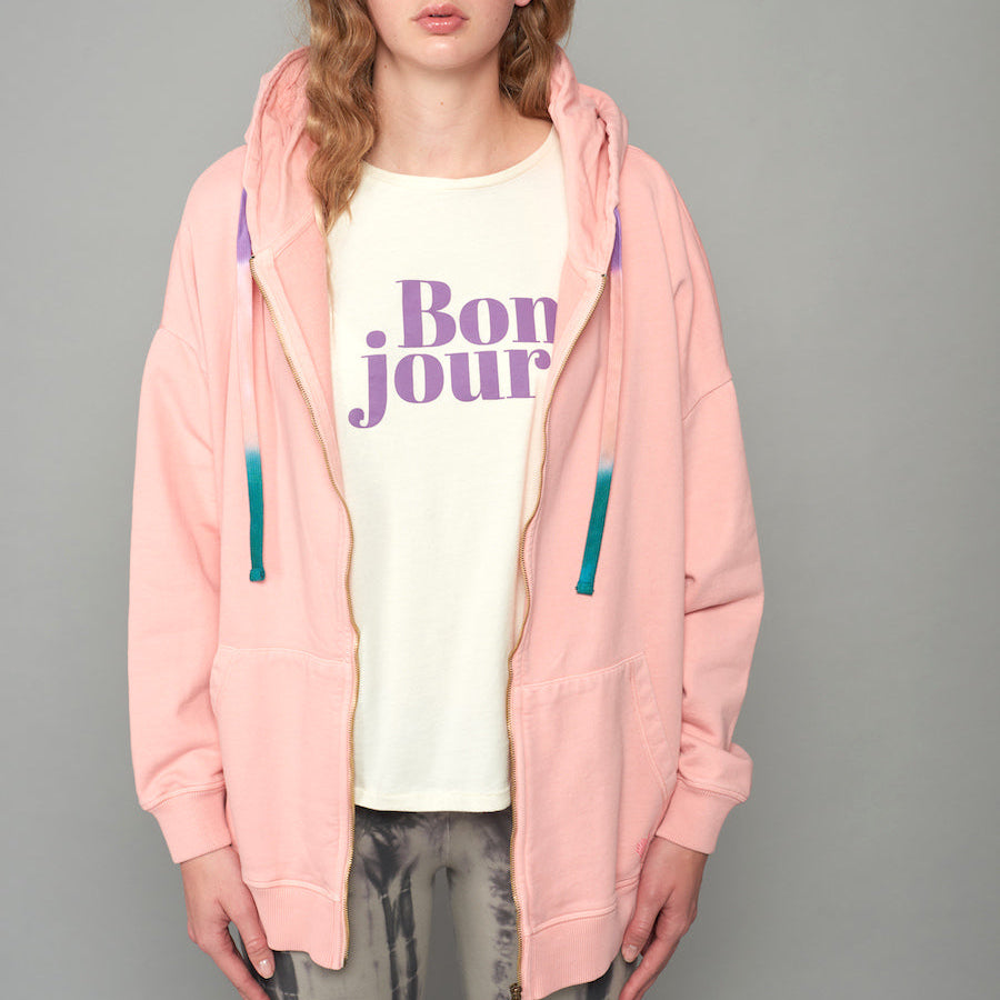 Person trägt ein weißes T-Shirt mit lila "Bonjour"-Schriftzug und eine rosa Kapuzenjacke mit blauen Kordeln.