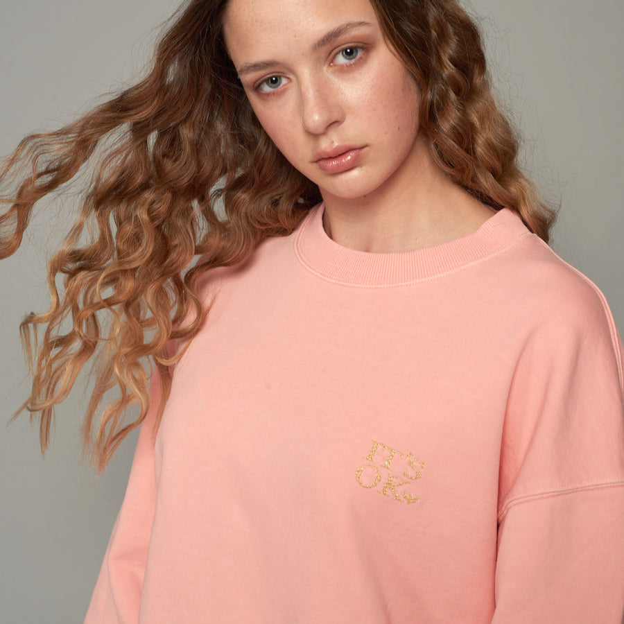 Eine Frau mit lockigem Haar trägt einen rosa Pullover mit der Aufschrift "It's OK".