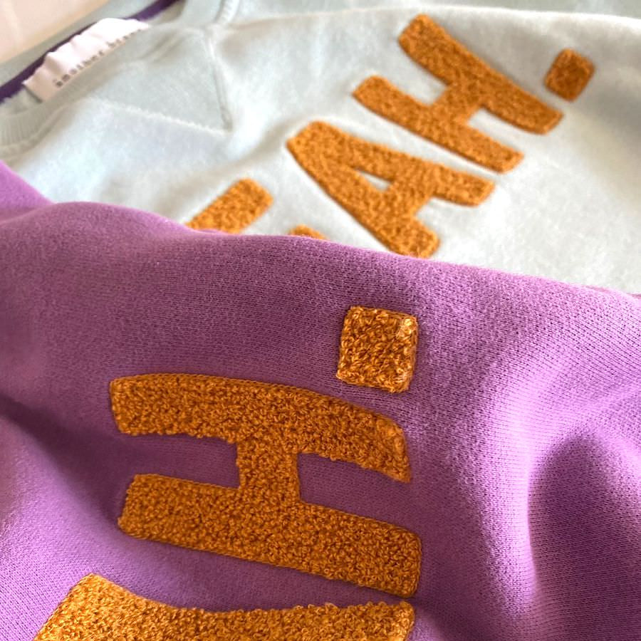 Zwei Pullover mit gestickten, gelb-orangen Texten auf lila und hellblauem Hintergrund.