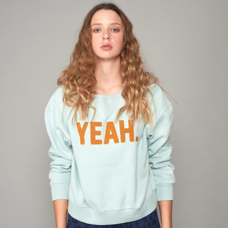 Frau mit langen, lockigen Haaren trägt einen mintgrünen Pullover mit „YEAH.“-Aufdruck und schaut geradeaus.