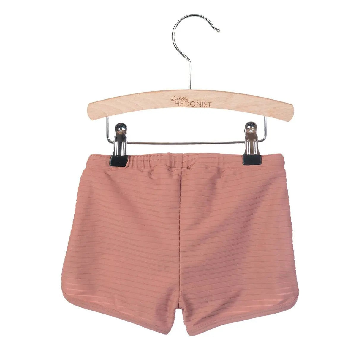 Hellrosa gerippte Shorts für Kinder, die auf einem Holzbügel mit Metallklammern befestigt sind. Auf dem Bügel ist Little Hedonist eingraviert, und die Shorts haben einen elastischen Bund.