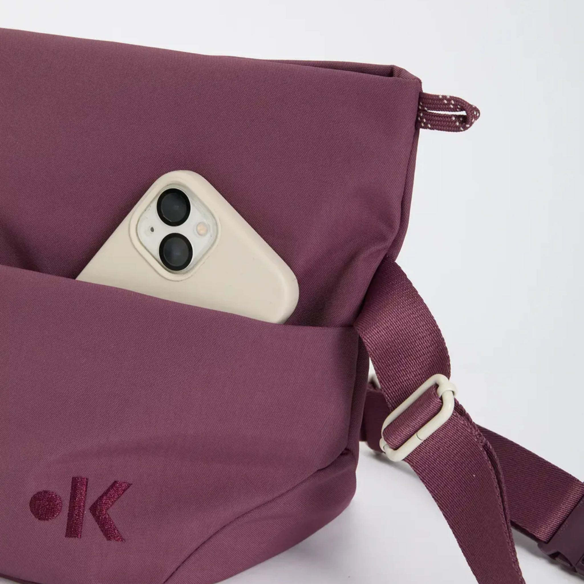 Mauvefarbene Crossbody-Tasche mit verstellbarem Riemen und einer Vordertasche, die ein weißes Smartphone mit zwei Kameras enthält. Die Tasche hat ein minimalistisches gesticktes Logo auf der Vorderseite.