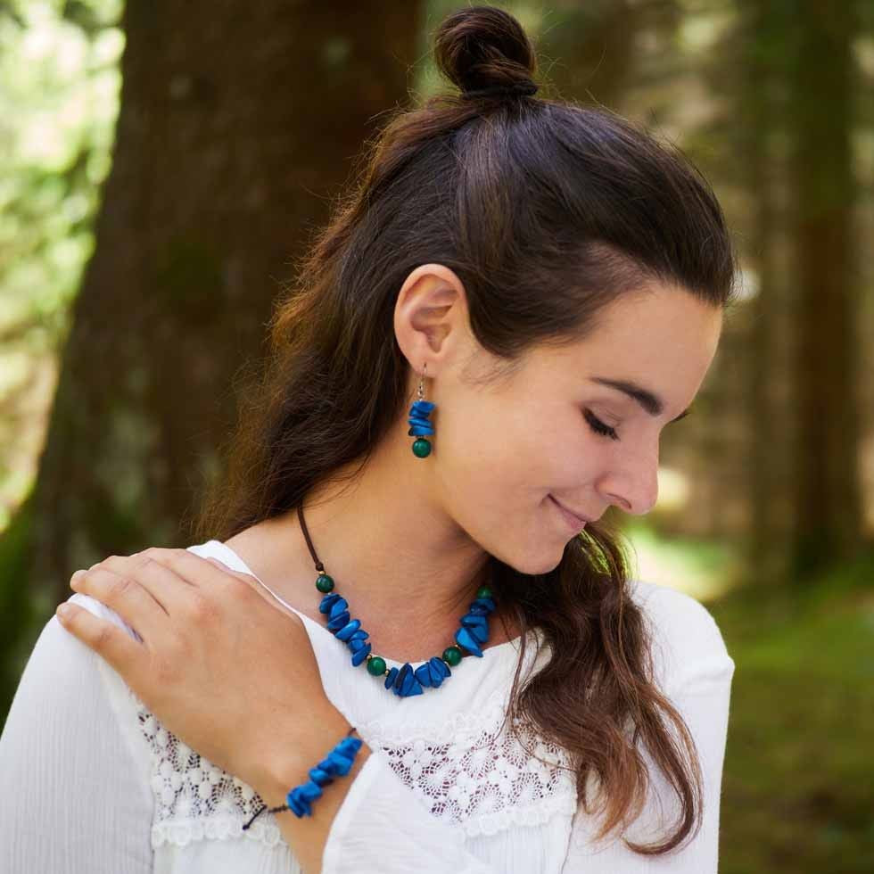 Eine Frau mit langen braunen Haaren, weißer Bluse und blauem Steinschmuck (Halskette, Ohrringe, Armband) blickt mit einem leichten Lächeln auf eine sonnenbeschienene Waldlandschaft herab.