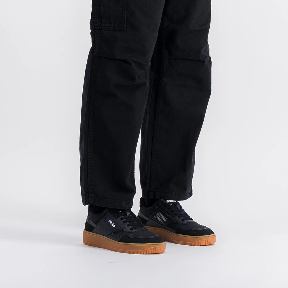 Person in schwarzer Hose und schwarzen Veja-Turnschuhen mit hellbraunen Sohlen, stehend vor einem schlichten weißen Hintergrund. Das Bild ist so beschnitten, dass nur die Beine und Füße zu sehen sind.