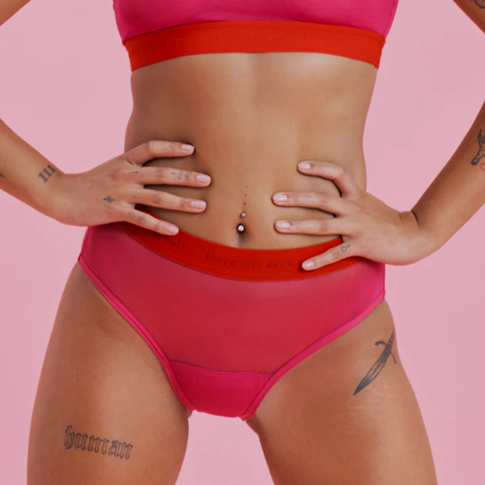 Person im pinken Unterwäsche-Set mit Tattoos auf der Haut vor rosa Hintergrund, die Hände auf den Hüften.