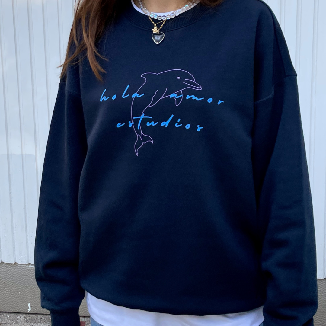 Person, die ein marineblaues Sweatshirt mit dem Umriss eines Delphins und der Aufschrift hola amor estudios in blauer Schrift trägt, steht vor einer hellen Wand.