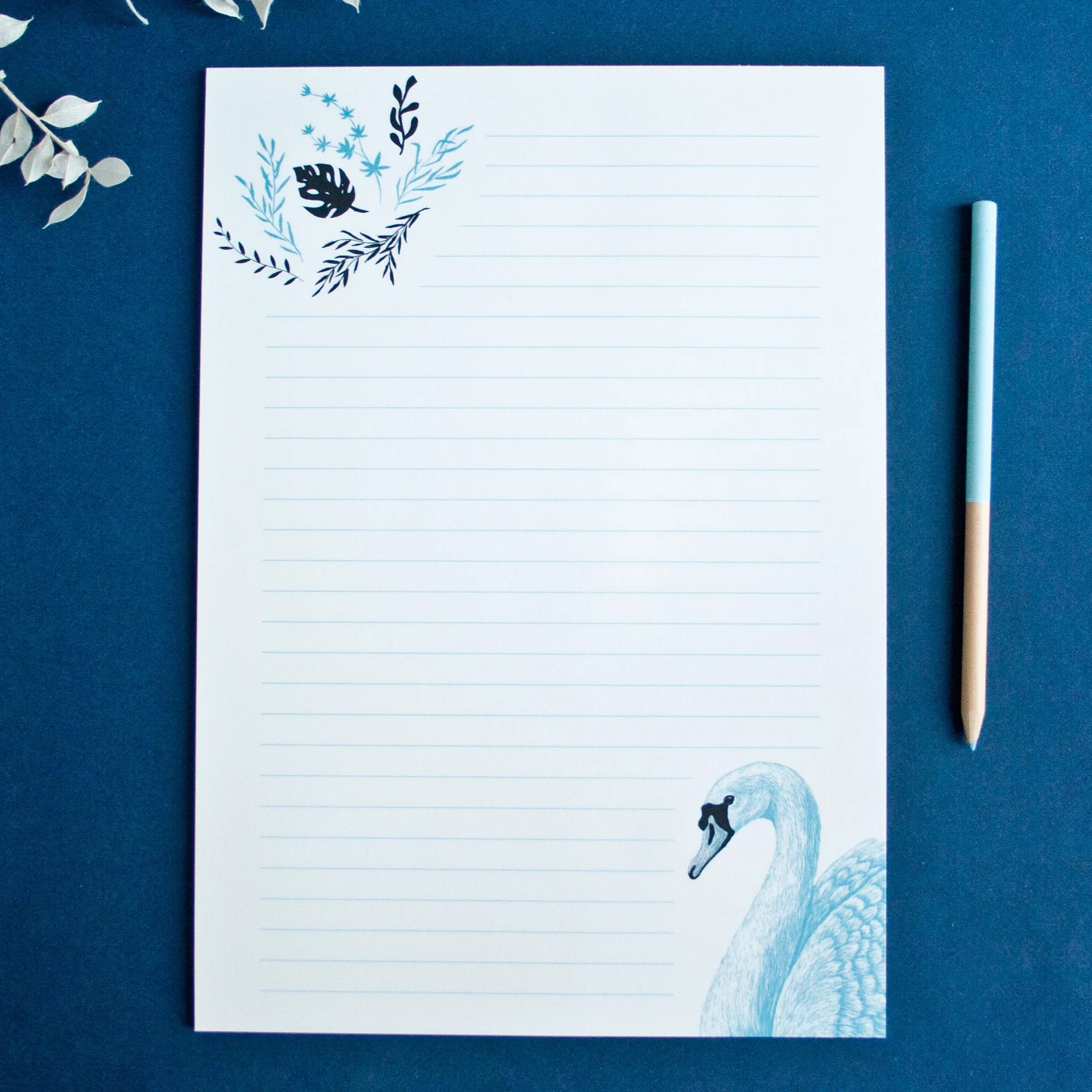Blauer Notizblock mit Pflanzen- und Schwanillustrationen auf einem blauen Tisch neben einem passenden Stift.