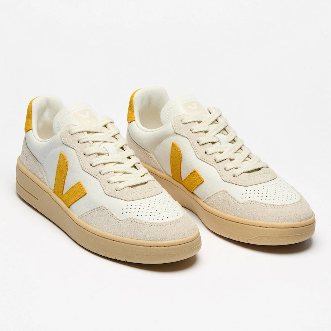 Ein Paar weißer Turnschuhe mit beigen Sohlen und gelben V-förmigen Logos an den Seiten, die auf einem einfarbig weißen Hintergrund abgebildet sind.