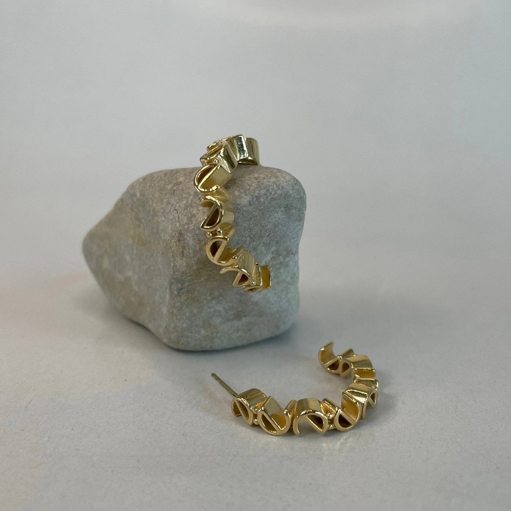 Goldene Creolen mit gewelltem Design, eine lehnt an einem grauen Stein, die andere liegt auf einer hellen Oberfläche.