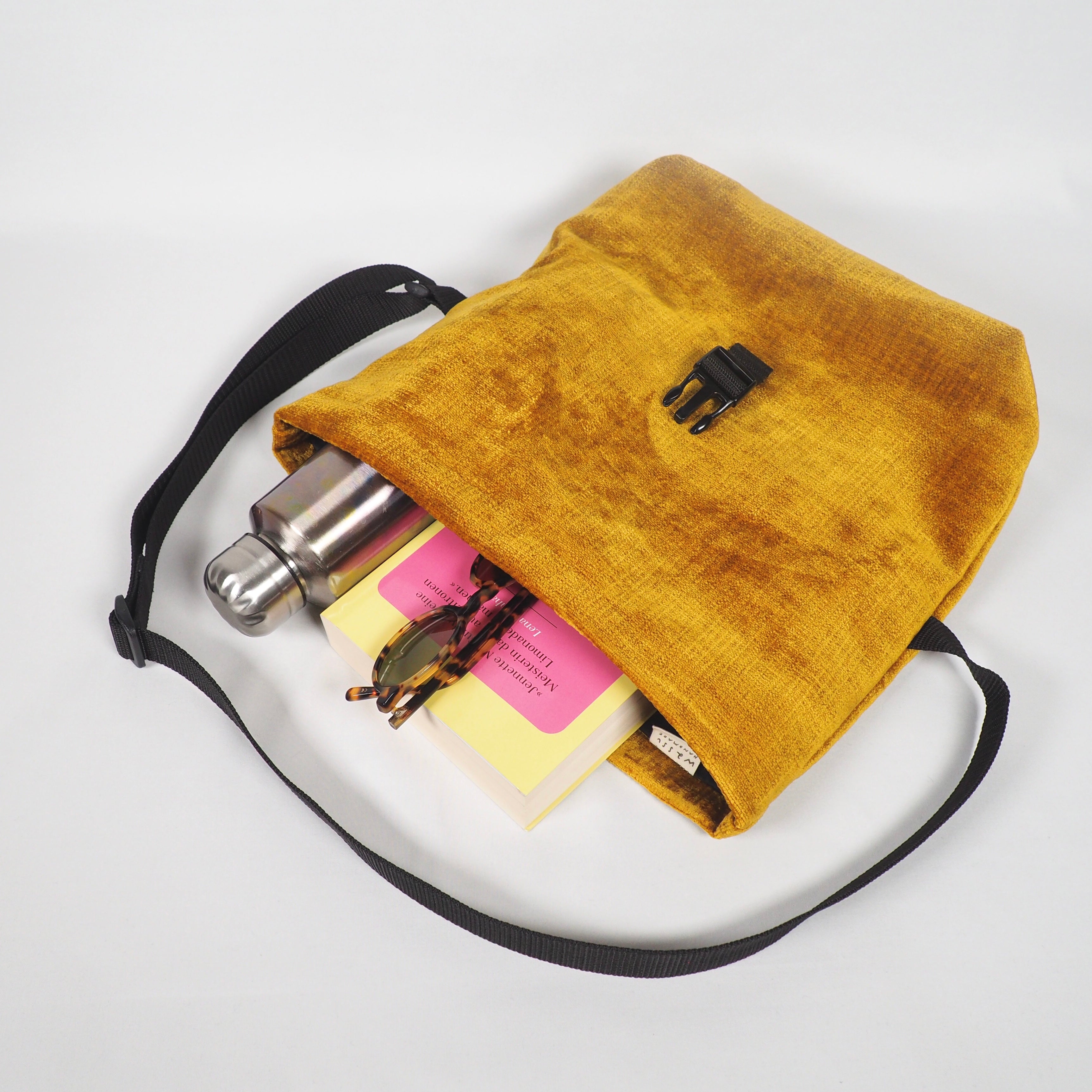 Offene gelbe Tasche mit einer Thermosflasche, Büchern und einer Sonnenbrille darin auf weißem Hintergrund.