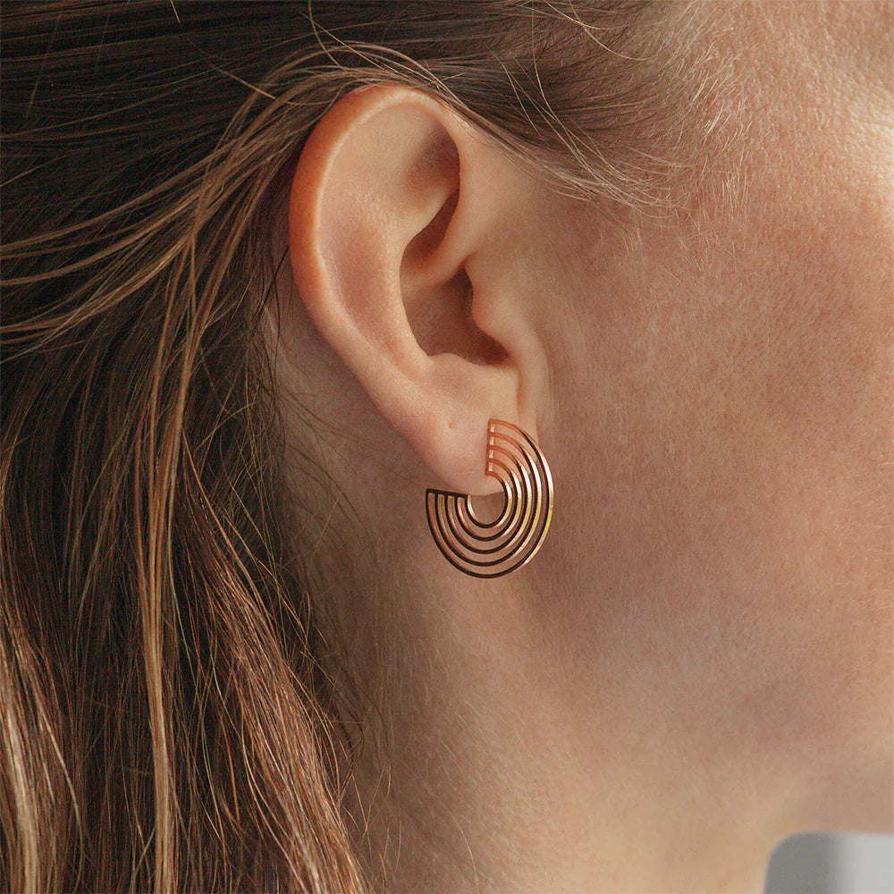 Nahaufnahme eines Frauenohrs, das einen modernen, halbrunden Goldohrring mit konzentrischen Bögen trägt. Ihr Haar ist hinter ihr Ohr gesteckt, und der Hintergrund ist leicht unscharf.