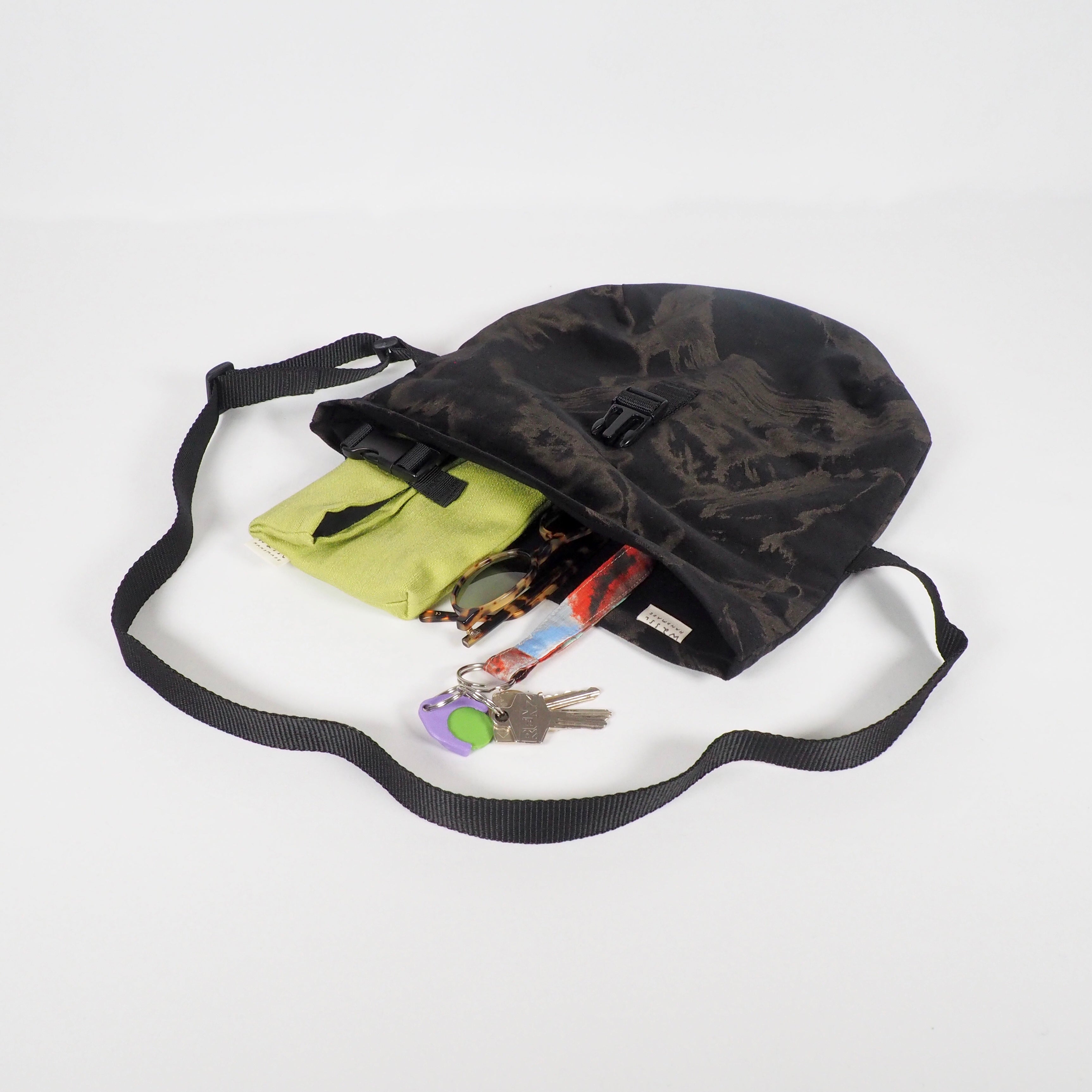 Schwarze Umhängetasche mit offenem Fach, gefüllt mit einem grünen Etui, Sonnenbrille, buntem Taschentuch und Schlüsseln.