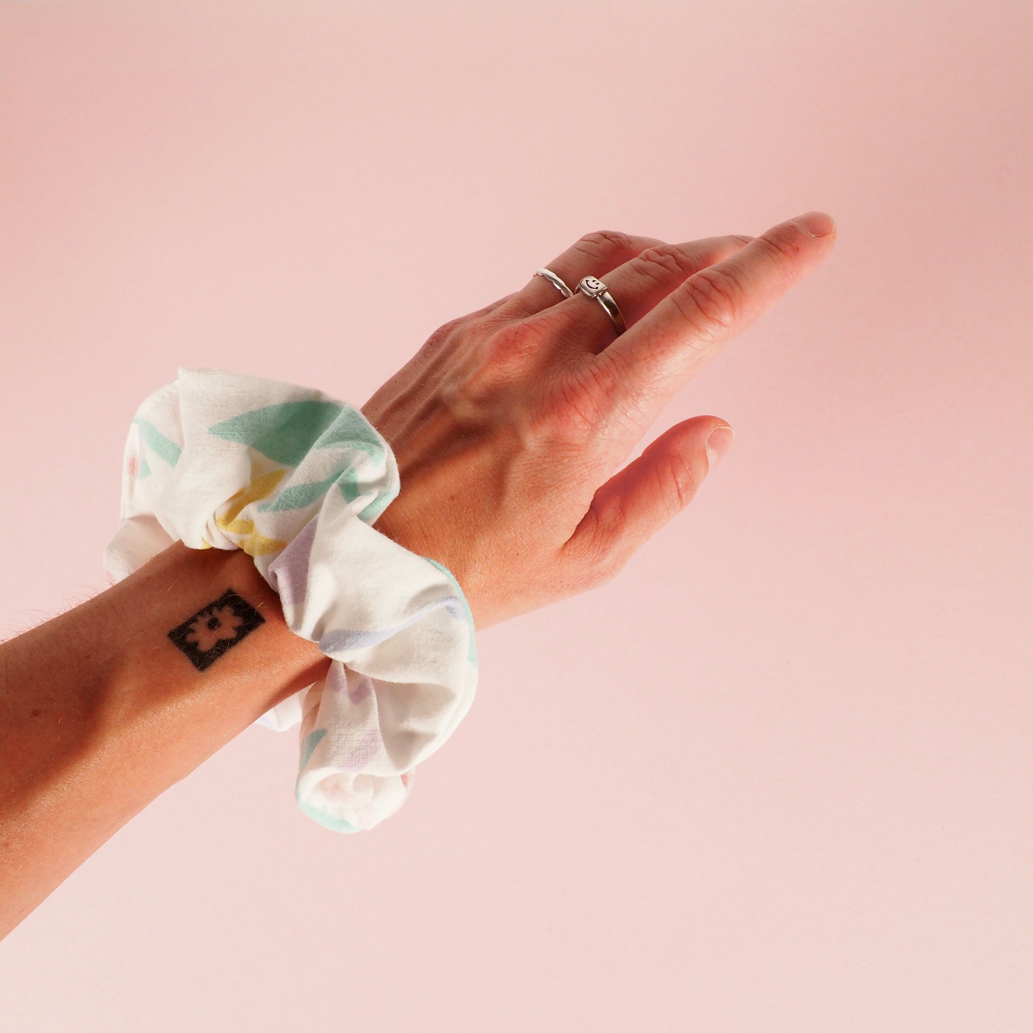 Hand mit Tattoo und pastellfarbenen Haarband, vor rosa Hintergrund.
