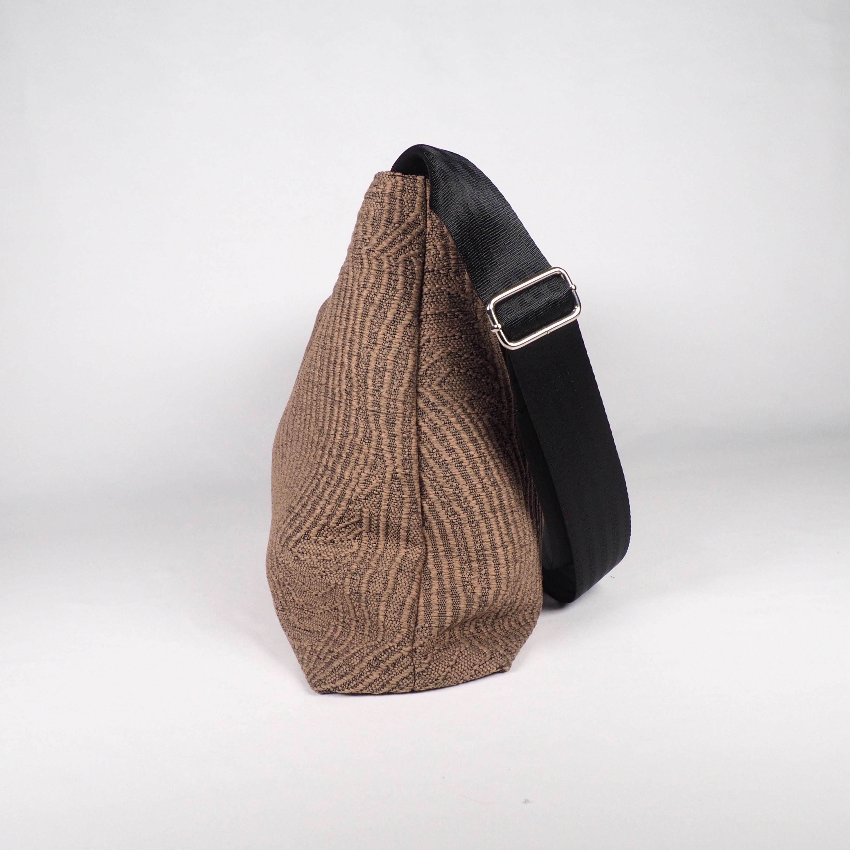 Braune, strukturierte Stofftasche mit breitem, schwarzem Schulterriemen und silberner Schnalle, seitlich fotografiert.