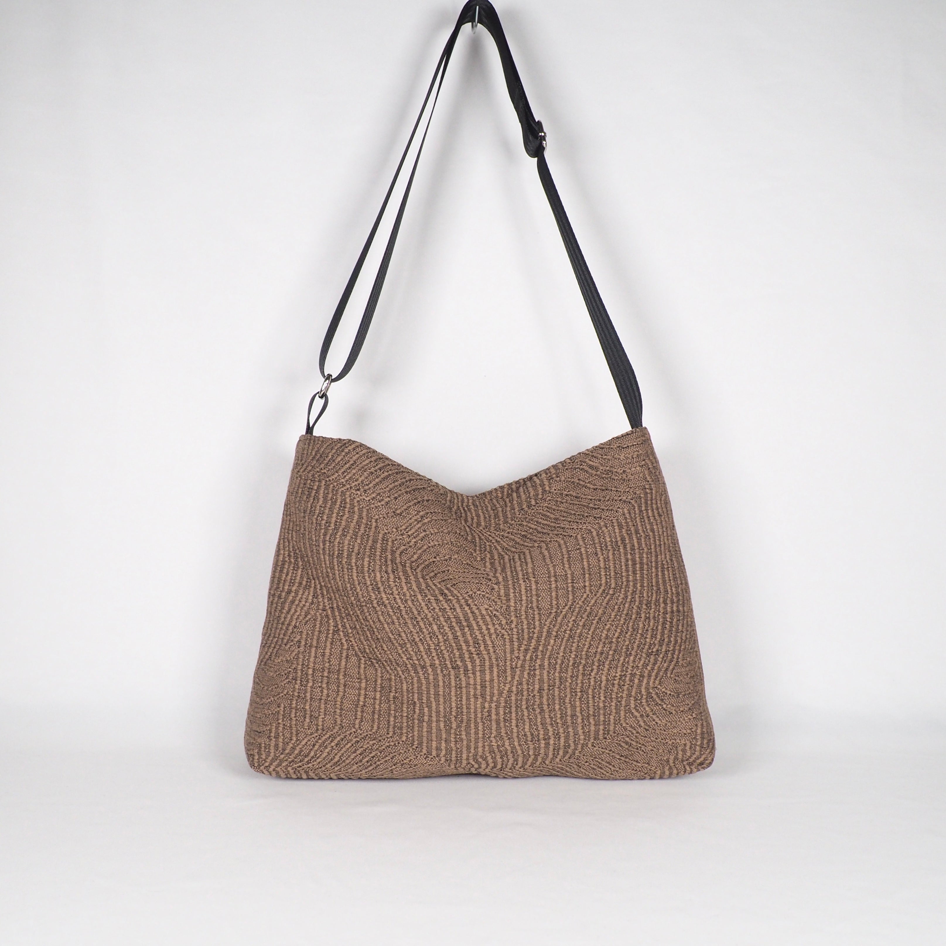 Braune Stofftasche mit schwarzem Schulterriemen, vor weißem Hintergrund platziert.