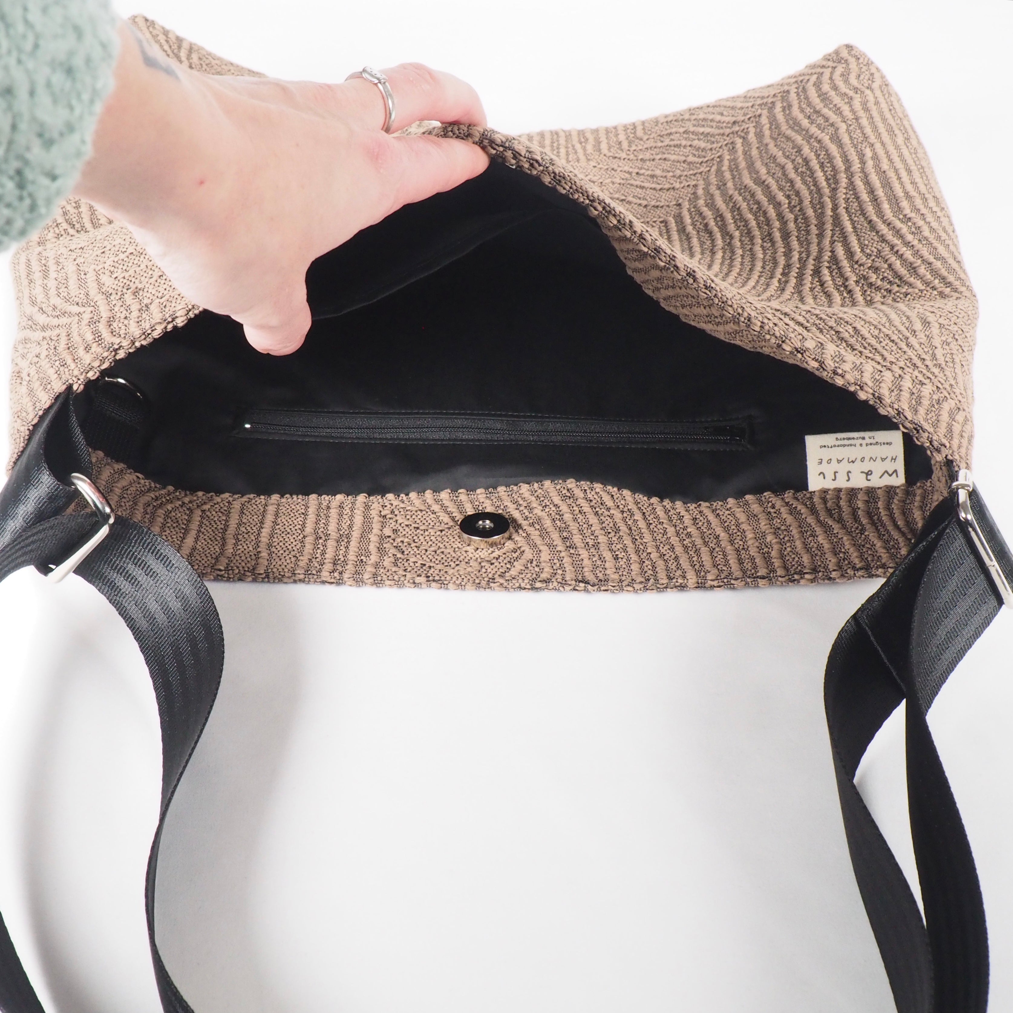 Geöffnete braune Tasche mit schwarzem Reißverschlussfach und Etikett. Eine Hand hält die Tasche geöffnet.