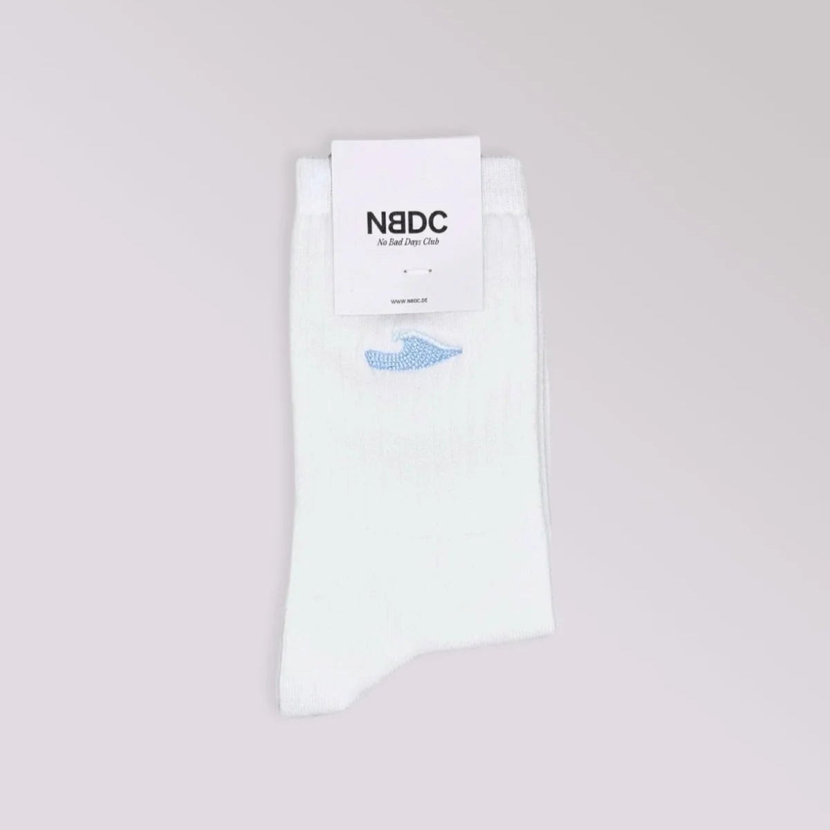 Ein Paar weiße Socken, ordentlich gefaltet, mit einem kleinen blauen gestickten Detail, das an einem weißen Etikett mit der Aufschrift N8DC Just a Joy Club auf hellgrauem Hintergrund befestigt ist.