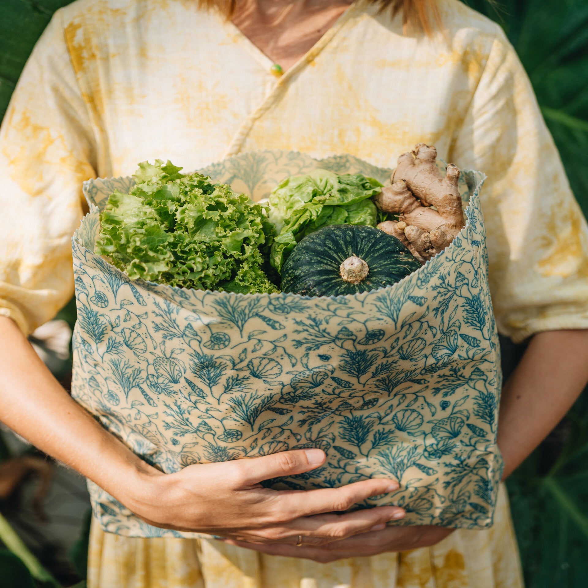 Eine Person in einem gelben Kleid hält eine wiederverwendbare gemusterte Tasche, die mit frischem Gemüse gefüllt ist, darunter Salat, ein grüner Kürbis und Ingwer. Im Hintergrund ist grünes Laub zu sehen.