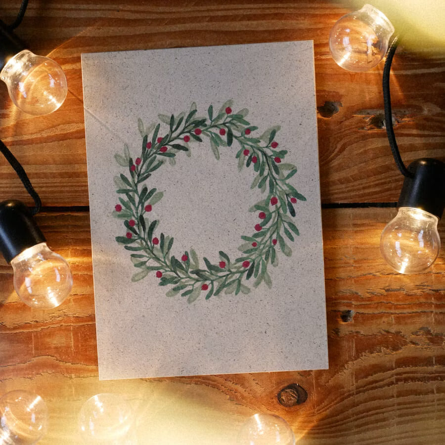 Karte mit gemaltem Kranz aus grünen Blättern und roten Beeren, umgeben von Lichterketten auf einem Holztisch.