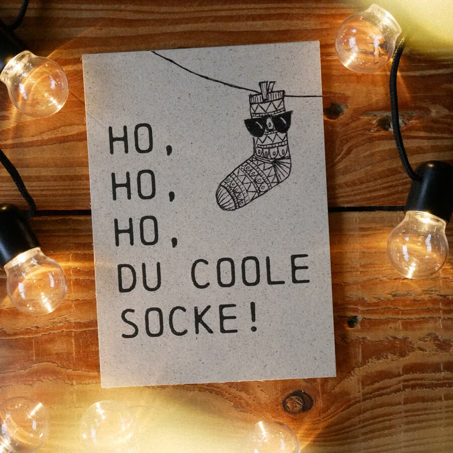Karte mit der Aufschrift "HO, HO, HO, DU COOLE SOCKE!" und einer illustrierten Socke, umgeben von Lichterketten.