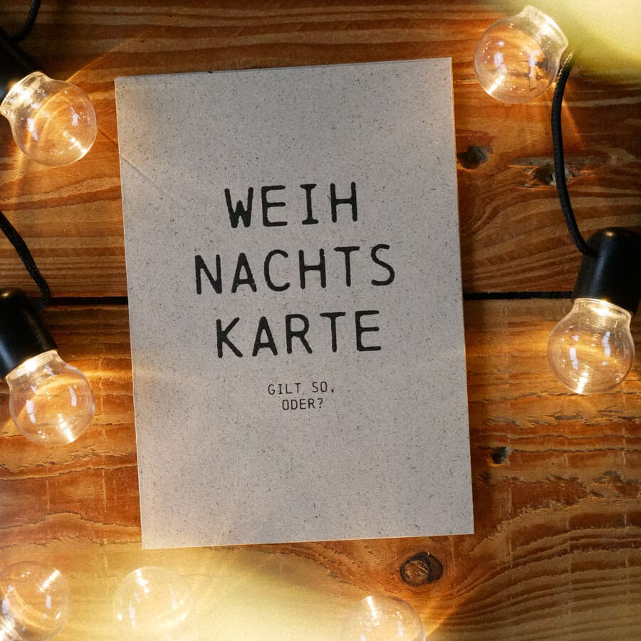 Karte mit der Aufschrift "WEIH NACHTS KARTE GILT SO, ODER?" auf Holztisch, umgeben von Lichterketten.