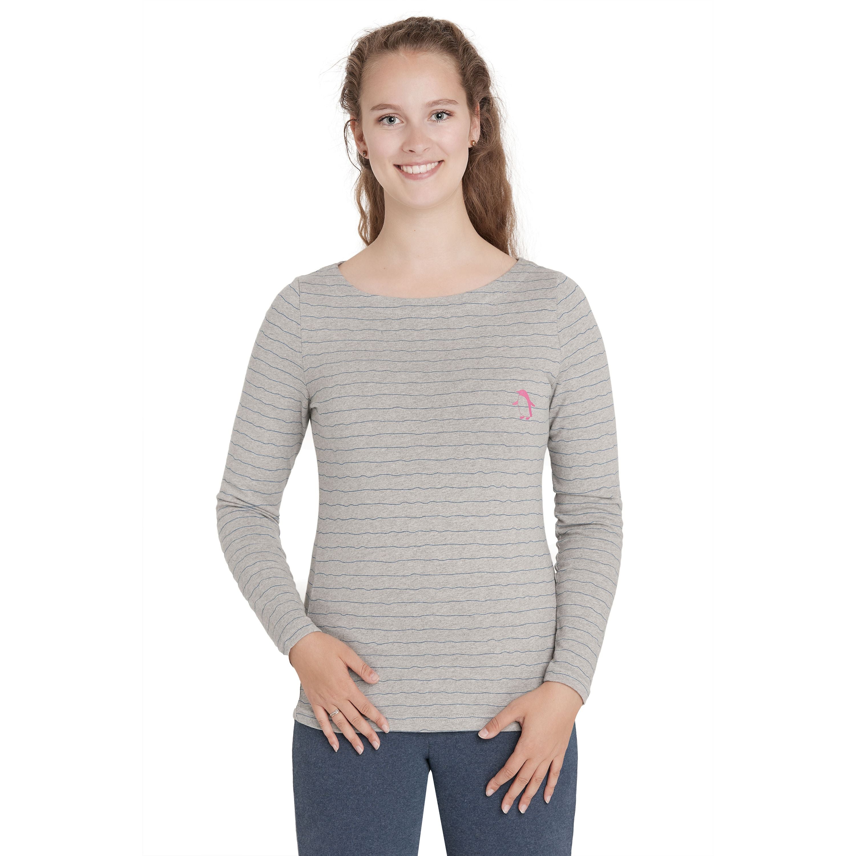 Eine junge Frau mit langen braunen Haaren steht lächelnd da, trägt ein hellgraues, langärmeliges, gestreiftes Hemd und eine dunkelblaue Hose und hat die Hände in die Hüften gestemmt. Der Hintergrund ist einfarbig weiß.