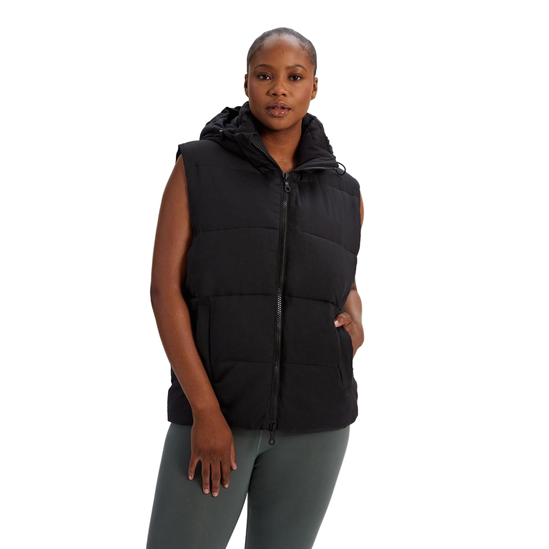 Eine Frau, die eine schwarze, ärmellose Pufferweste mit Kapuze und Frontreißverschluss trägt, steht vor einem schlichten weißen Hintergrund. Sie hat eine Hand in der Tasche und ist mit dunkelgrünen Leggings bekleidet.