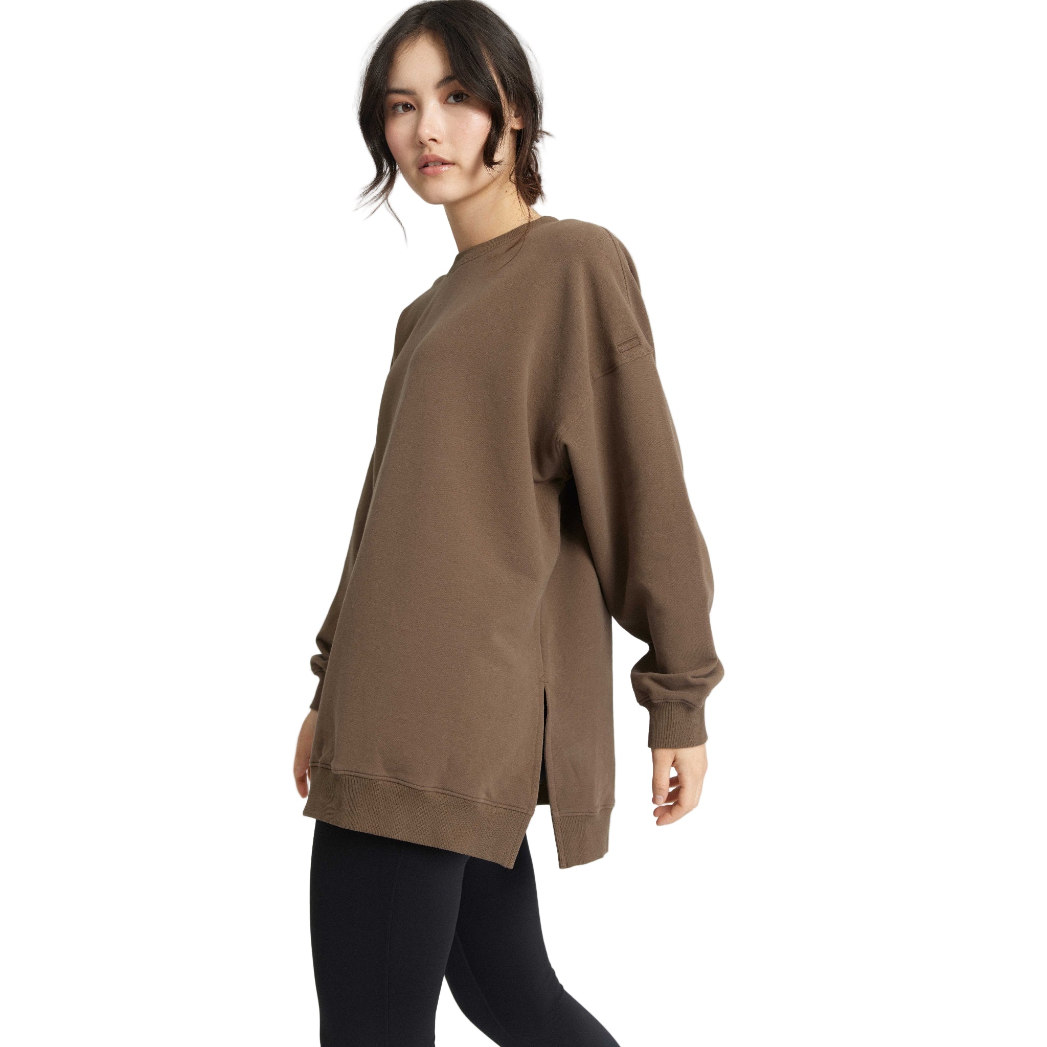 Eine Frau mit dunklem Haar in einem lockeren Pferdeschwanz trägt ein übergroßes braunes Sweatshirt mit Seitenschlitzen und schwarze Leggings. Sie steht seitlich und schaut in die Kamera vor einem weißen Hintergrund.