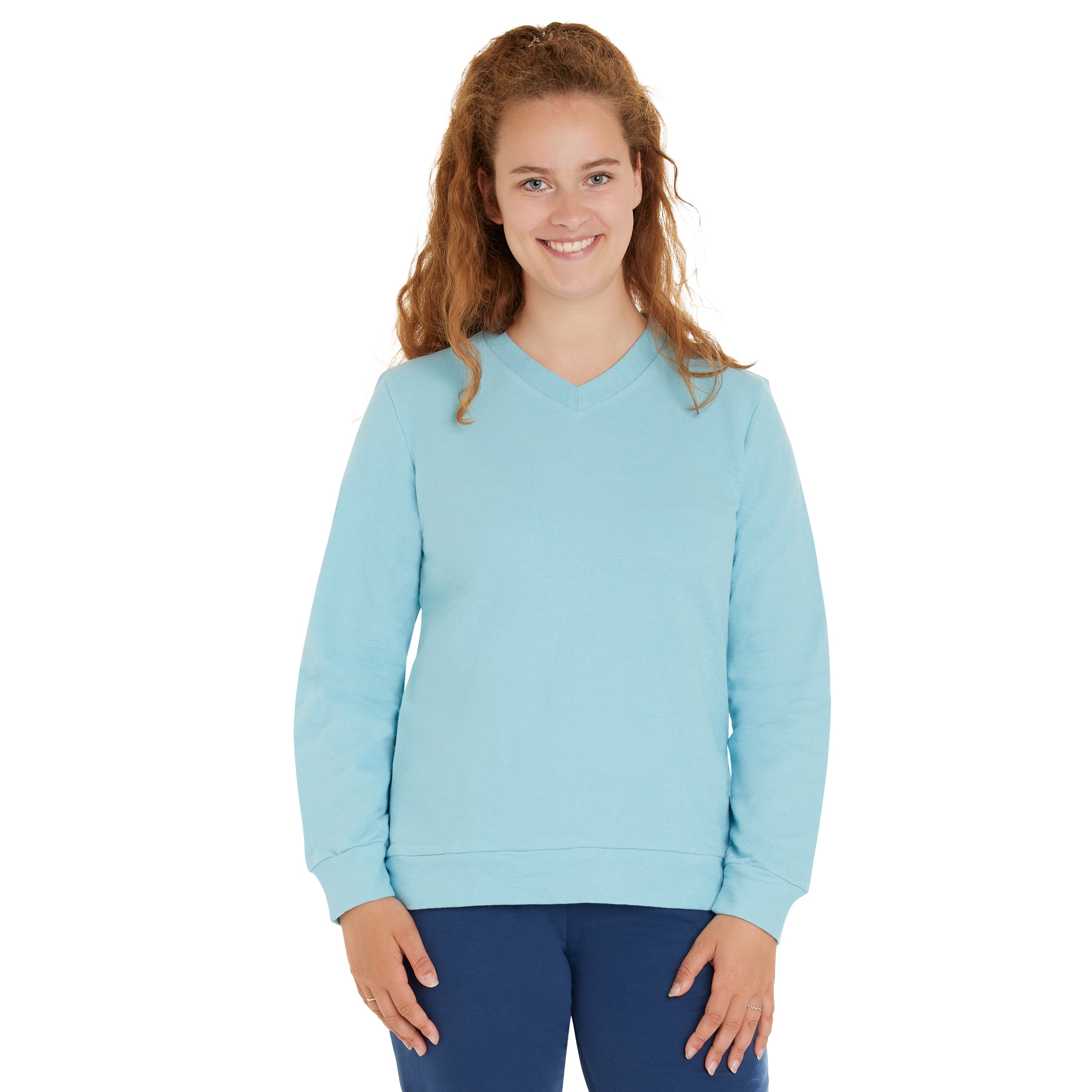 Eine Frau mit langen lockigen Haaren, die ein hellblaues Sweatshirt mit V-Ausschnitt und eine dunkelblaue Hose trägt, steht lächelnd vor einem schlichten weißen Hintergrund.