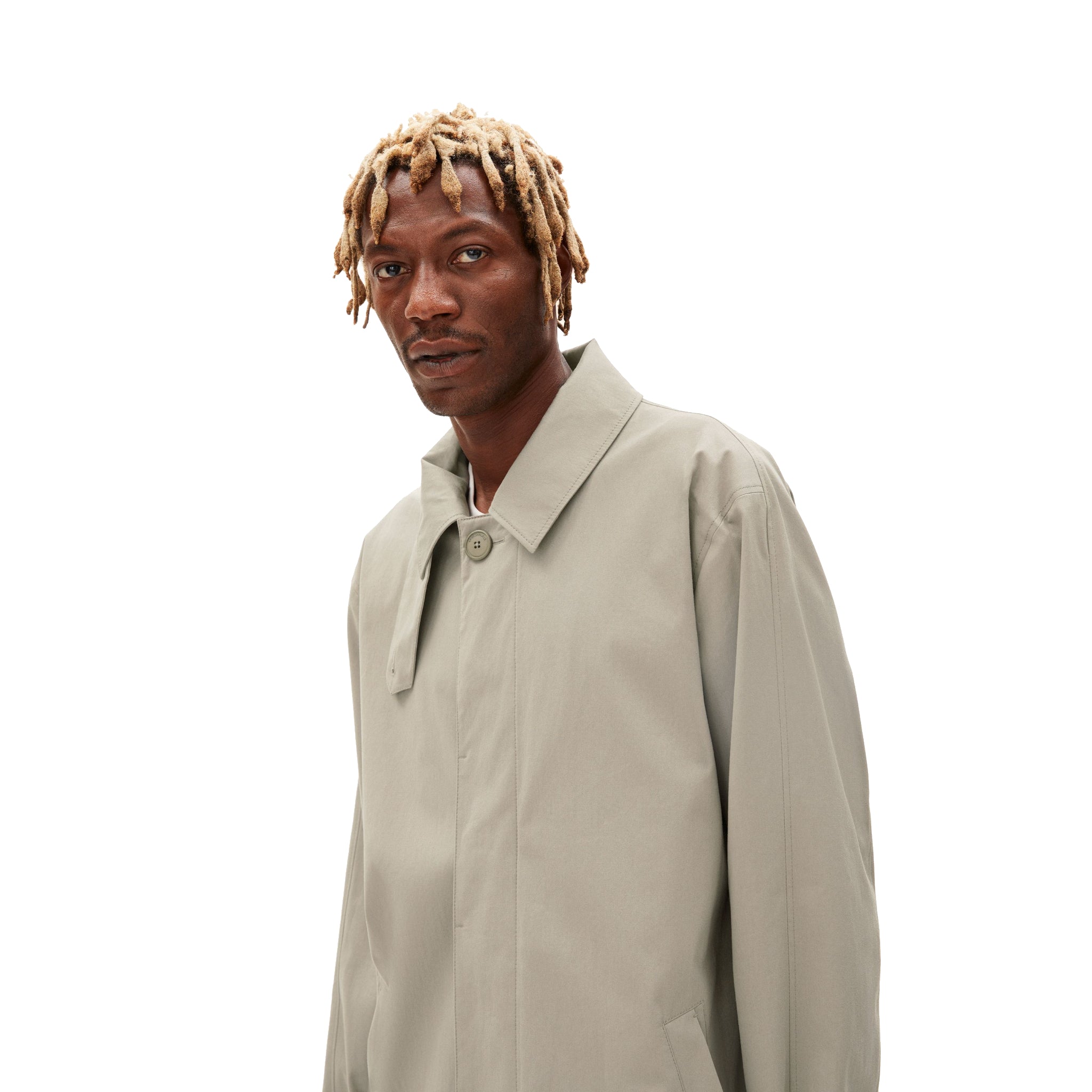 Ein Mann mit blondem Dreadlocks trägt einen beigen Mantel vor weißem Hintergrund.