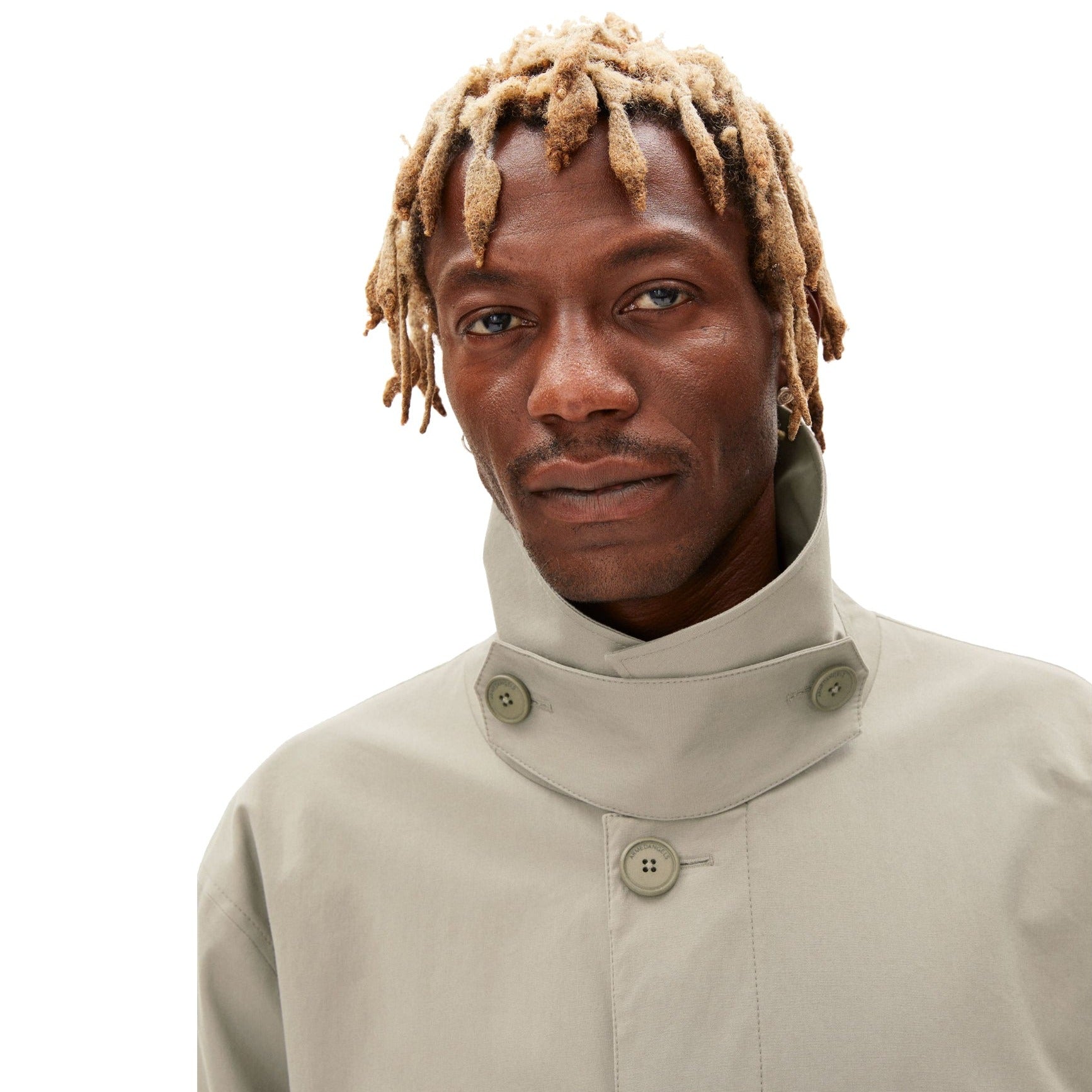 Mann mit blonden Dreadlocks trägt einen beigefarbenen Mantel mit hohem Kragen und Knöpfen vor weißem Hintergrund.