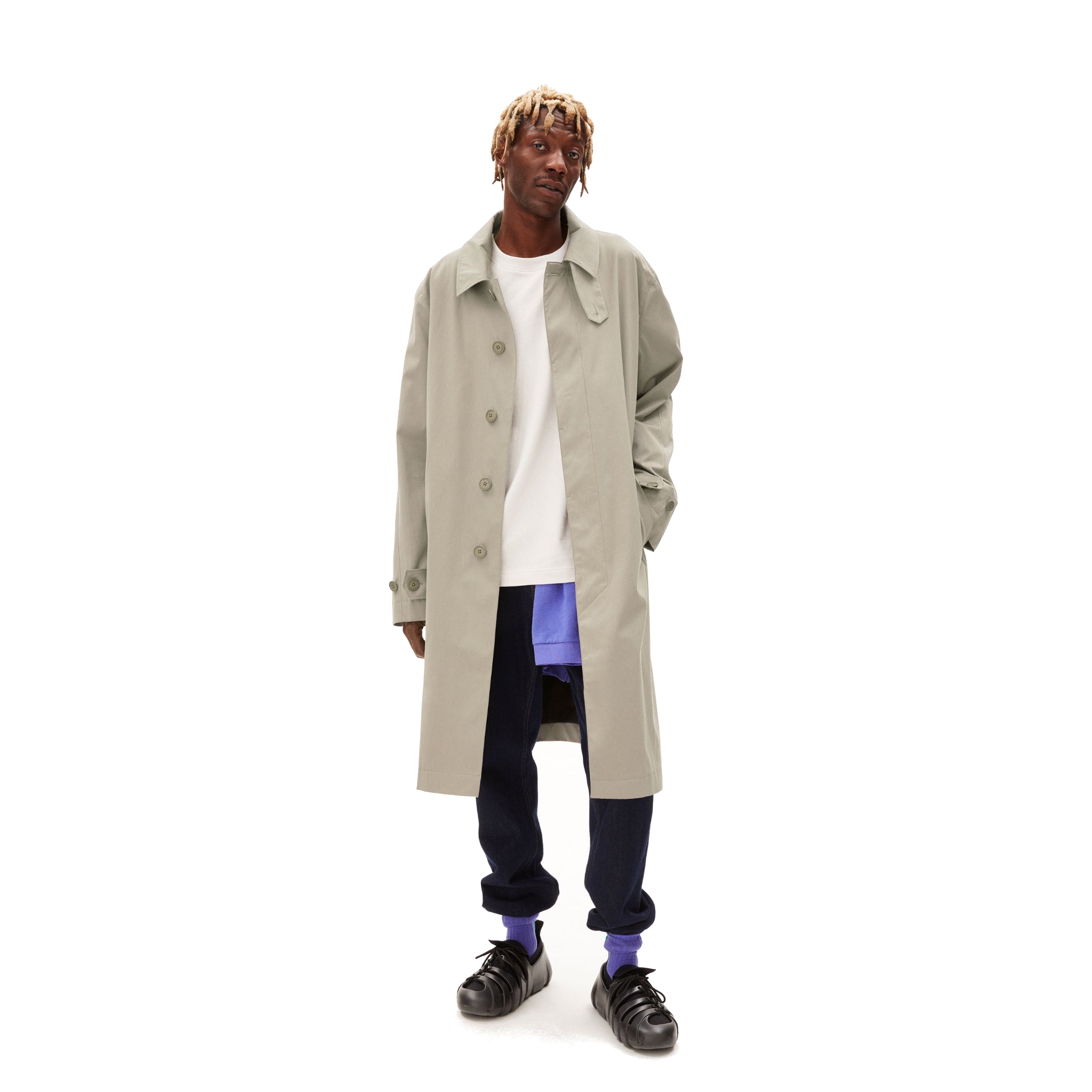 Eine Person trägt einen langen grauen Mantel über einem weißen Shirt, blauen Hosen und schwarzen Schuhen, vor weißem Hintergrund.