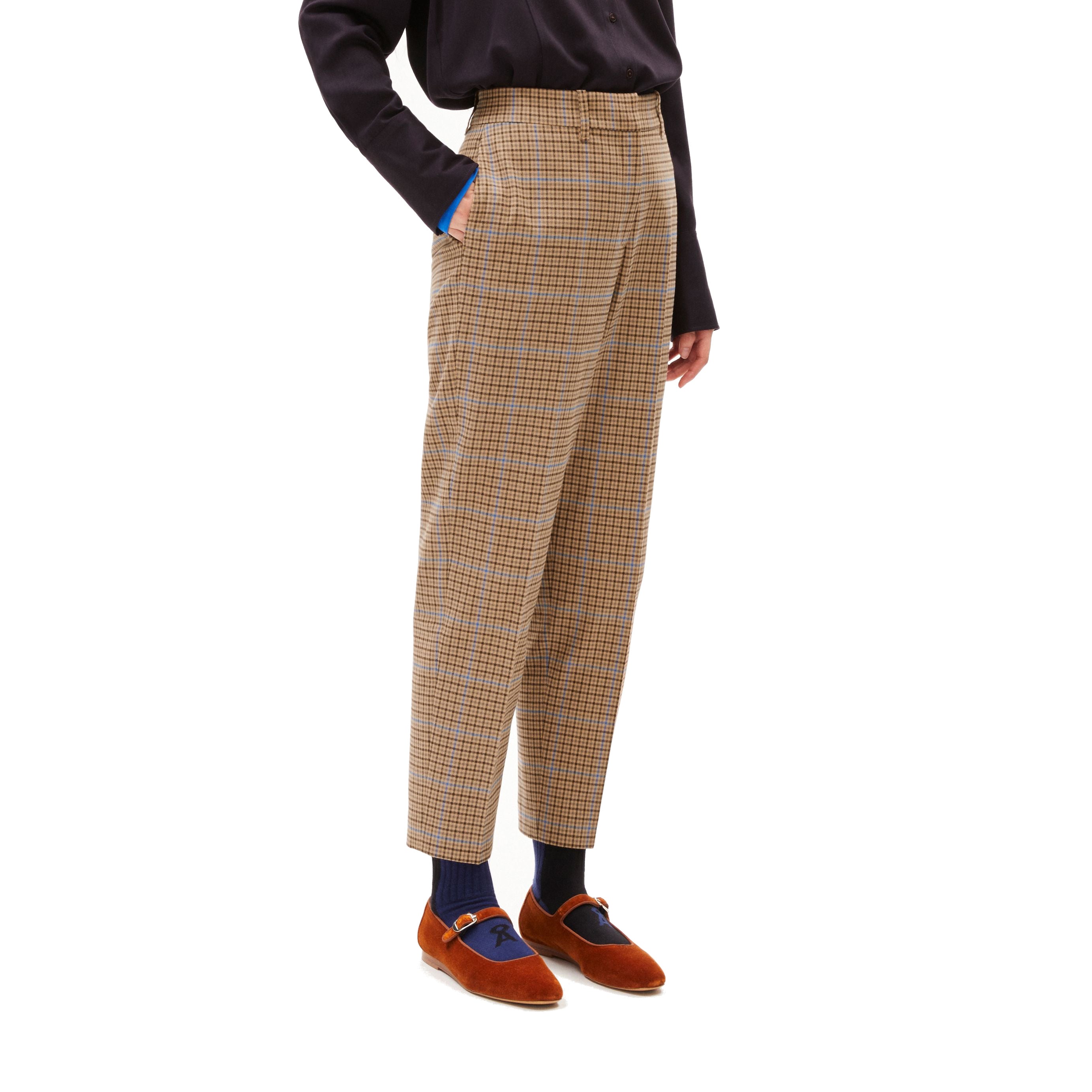 Person in karierter Hose und braunem Hemd, trägt orangefarbene Schuhe und blaue Socken, Hände in den Hosentaschen.