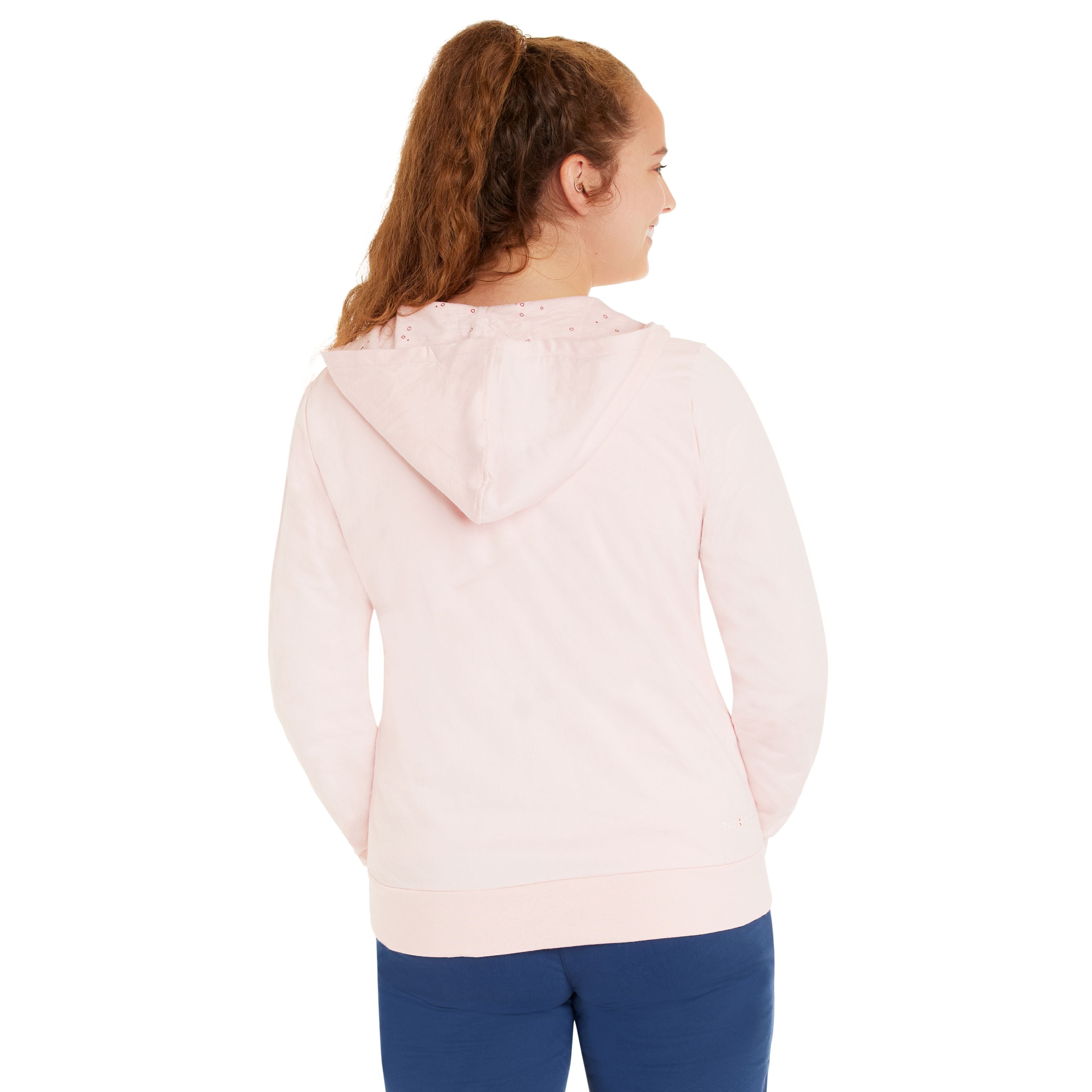 Eine Frau mit langen braunen Haaren in einem Pferdeschwanz trägt einen hellrosa Hoodie und eine blaue Hose. Sie steht mit den Händen in den Taschen ihres Kapuzenpullis und schaut von der Kamera weg.